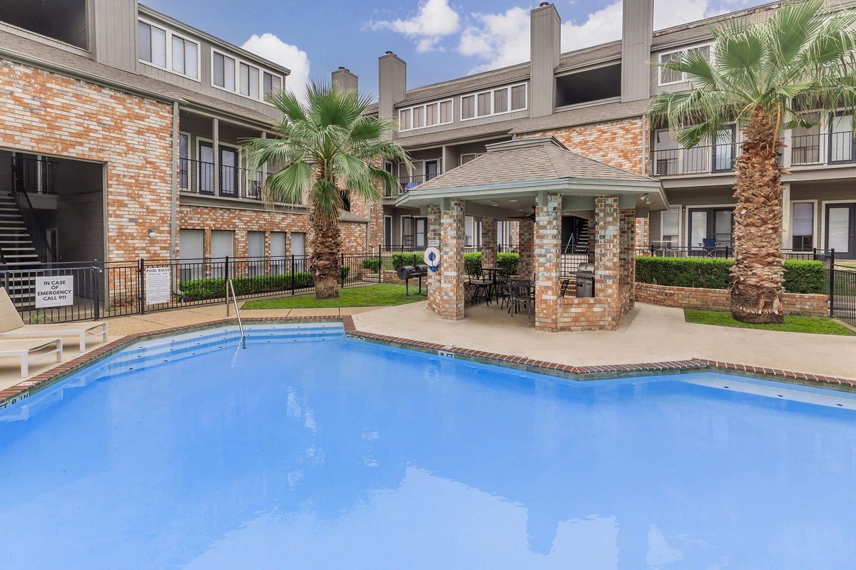 A well-maintained swimming pool surrounded by palm trees, with a gazebo featuring tables and chairs. The pool area is located within an apartment community, which is visible in the background with brick exterior and greenery. The sky is partly cloudy, adding to the inviting atmosphere.