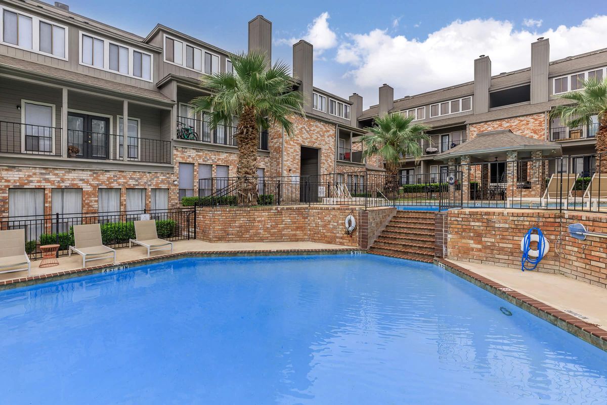 A sunny view of a residential pool area featuring a clear blue pool surrounded by palm trees and lounge chairs. The pool is located at the base of a multi-story apartment building with brick and wood exteriors. There are staircases leading into the pool area, along with lifebuoys for safety.