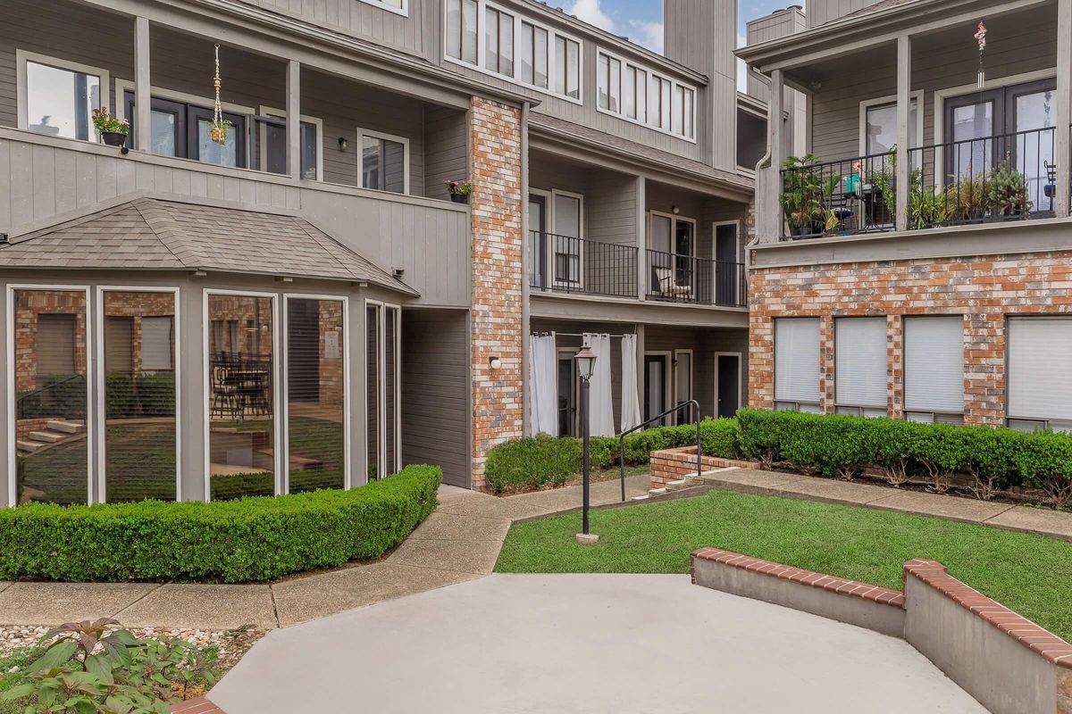 A courtyard view of a multi-story apartment building featuring gray and red brick facades. The area includes well-maintained greenery, a walking path, and a gazebo-like structure. Several balconies are visible with potted plants. The setting is bright and open, suggesting a pleasant outdoor living space.