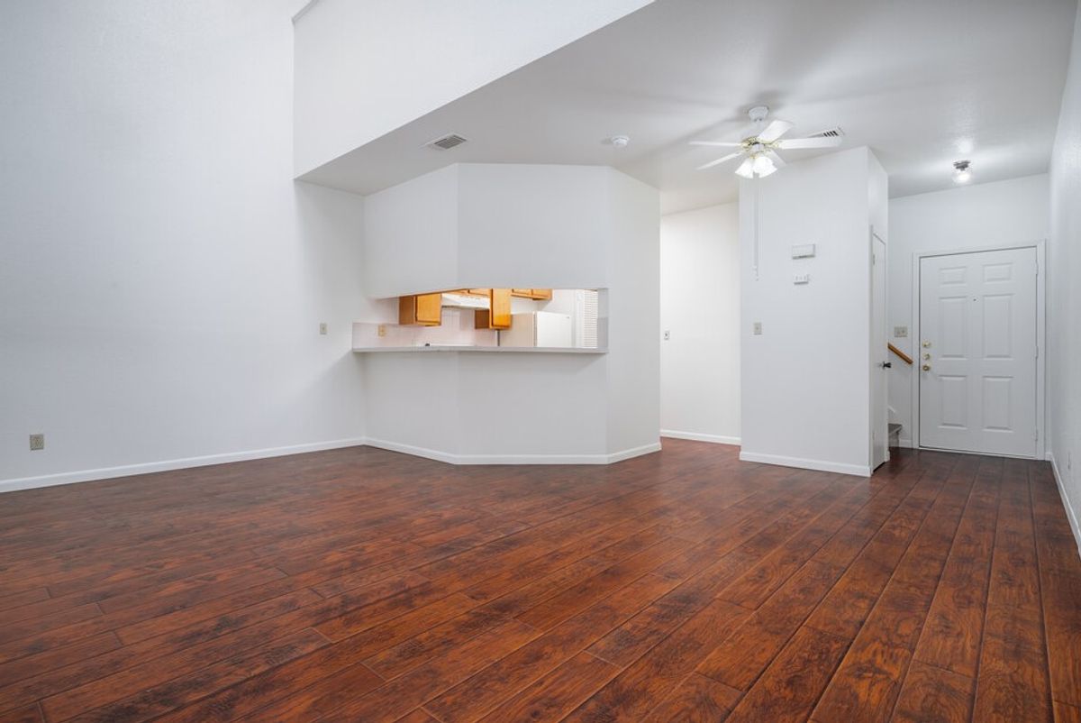 Empty living room featuring light-colored walls and dark wooden flooring. A small kitchenette is visible in the background with open shelving. The space is bright, with a ceiling fan and an entry door on the right.