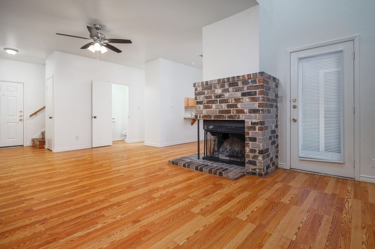 A spacious interior of a living room featuring a brick fireplace, hardwood flooring, a ceiling fan, and neutral-colored walls. A front door and a side door with a window are visible, alongside an open doorway leading to another room. The overall ambiance is bright and inviting.