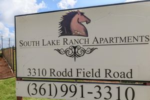 Sign for South Lake Ranch Apartments featuring a horse graphic. The text includes the address "3310 Rodd Field Road" and a phone number "(361) 991-3310." The background shows a partly cloudy sky, and the sign is positioned at the entrance to the apartment community.