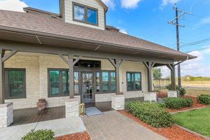 A welcoming entrance of a modern building featuring a stone façade, large windows, and a covered porch. Lush green shrubs and mulch border the walkway, which leads to a glass door. A clear sky with scattered clouds is visible in the background. Utility poles line the road nearby.