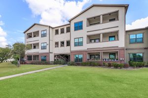 A three-story apartment building with multiple balconies and large windows, situated in a well-kept grassy area. The exterior features a mix of light and dark siding, with potted plants visible on the ground floor balcony. The surrounding lawn is neatly maintained, and the sky above is partly cloudy.
