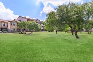 A sunny outdoor scene featuring well-manicured green lawns and several trees. In the background, there are two multi-story residential buildings with a modern design. Fluffy white clouds are scattered across a blue sky, enhancing the bright and inviting atmosphere of the area.