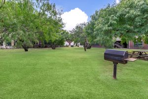 A spacious green backyard featuring a well-maintained lawn with several trees and a picnic table in the distance. A black barbecue grill is positioned prominently in the foreground, surrounded by lush greenery under a bright blue sky with fluffy white clouds.