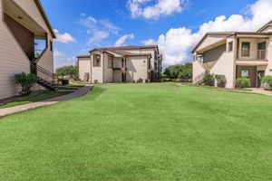 A spacious green lawn surrounded by two-story apartment buildings under a blue sky with fluffy clouds. The pathway leading through the grass enhances the inviting atmosphere of the area. Lush shrubs and trees are visible, adding to the greenery of the landscape.