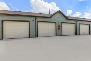 Row of four beige garage doors on a green building, set against a bright blue sky with fluffy white clouds. The concrete ground is smooth, and a red fire extinguisher is mounted beside the rightmost garage door. The setting appears well-maintained and modern.
