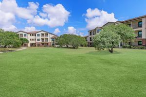 A lush green lawn surrounded by modern residential buildings under a bright blue sky with fluffy white clouds. Trees are dotted throughout the grassy area, creating a serene and inviting atmosphere. The scene conveys a peaceful community setting ideal for outdoor activities or relaxation.