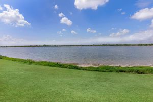 A serene landscape featuring a calm body of water bordered by lush green grass. Puffy white clouds float in a blue sky, with trees visible in the distance along the shore. The scene captures a peaceful, natural setting ideal for relaxation or outdoor activities.