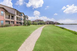 A peaceful residential area along a well-maintained path by a serene lake. Two-story apartment buildings with a mix of colors line the grassy shoreline. Blue skies with fluffy clouds create a calming atmosphere, ideal for leisurely walks or relaxing by the water.