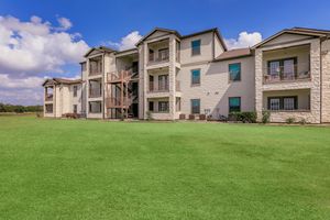 A multi-story apartment building with a light-colored facade and stone accents, surrounded by lush green grass and blue sky with fluffy clouds. The building features balconies with wooden railings and some visible decorative items, providing a modern residential aesthetic.