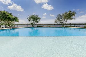 A serene swimming pool with clear blue water, surrounded by lush greenery and two trees on either side. The pool reflects clouds in a bright blue sky, with a view of a calm body of water in the background and a black fence enclosing the area. Perfect for relaxation and leisure.