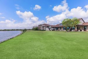 A scenic view of a grassy lawn beside a calm body of water, with a two-story building featuring large windows and a covered porch. The sky is filled with fluffy white clouds, creating a tranquil atmosphere. Perfect for relaxing or outdoor activities.