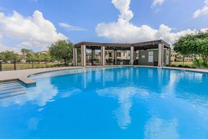 A large swimming pool with clear blue water, surrounded by a deck. In the background, there is a covered seating area and lush green trees. The sky is bright with fluffy white clouds, creating a relaxing outdoor atmosphere.