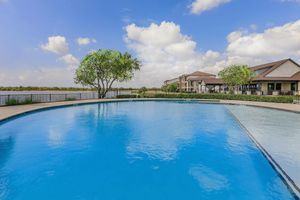 A tranquil swimming pool with clear blue water reflecting the sky, surrounded by lush greenery and trees. In the background, a scenic view of a lake is visible alongside elegant residential buildings and a vibrant landscape under a partly cloudy sky.