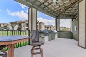 Outdoor grilling area featuring two BBQ grills under a wooden pavilion, with a brown table and chair nearby. The background includes multi-story residential buildings and a clear blue sky with fluffy white clouds. The setting is ideal for gatherings and leisure activities.