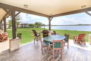 Outdoor seating area with wooden tables and chairs under a covered porch, overlooking a calm lake. Lush green grass surrounds the space, and a few trees are visible in the background alongside more buildings. The sky is partly cloudy, creating a serene and inviting atmosphere.