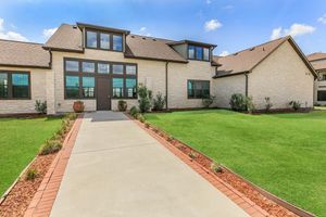A modern two-story house with large windows, light-colored stone exterior, and a brown roof. The front yard features a neatly manicured green lawn, bordered by a brick walkway and flower beds with small plants. The scene is set under a clear blue sky with a few clouds.
