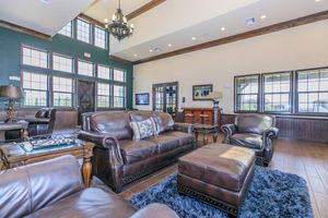 A spacious and elegantly furnished living room featuring leather sofas, a matching armchair, and a wooden coffee table on a plush area rug. Large windows allow natural light to fill the room, highlighting the stylish decor and warm wood accents on the walls and ceiling. A chandelier adds a touch of sophistication.