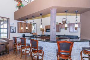 Modern kitchen with an open layout featuring dark wooden cabinets, a dark granite countertop, and multiple pendant lights hanging above. There are bar stools along the counter, and windows offering a view of the outdoors. Decor includes plants and decorative items on the shelves. Stylish and inviting atmosphere.
