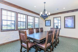 A spacious dining room featuring a large wooden table surrounded by eight high-backed chairs. The room has large windows with decorative frames, allowing natural light to flood in. A chandelier hangs from the ceiling, and artwork is displayed on the walls, enhancing the inviting atmosphere.