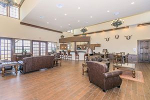 A spacious and elegantly designed lounge area featuring leather sofas, wooden tables, and chairs. Large windows let in natural light, and decorative chandeliers hang from the ceiling. The room has a warm color palette with accents like plants and wall-mounted decorative items, creating a welcoming atmosphere.