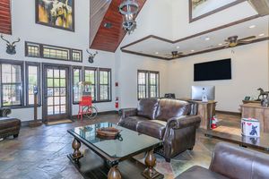 A spacious lobby area featuring a high ceiling with wooden beams, a large dark leather sofa, and a glass coffee table. Large windows with wooden slats provide natural light. A modern computer is on a desk, and decorative elements include wall art, an antique popcorn machine, and a fire extinguisher.