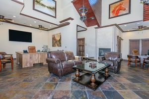 A spacious interior featuring leather armchairs and a glass coffee table on a slate floor. There’s a wooden desk in the background and artwork on the walls, including a painting of a horse. The room is well-lit with ceiling fans and a fireplace, creating a welcoming atmosphere.