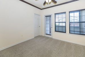 A spacious room with beige walls and a light brown ceiling. It features two large windows with black blinds and a door leading to a balcony. The floor is covered in beige carpet, creating a cozy atmosphere. The room appears well-lit and has a ceiling fan.
