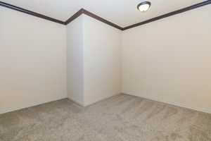 A vacant room with beige walls and plush beige carpet. The space features a corner with a simple ceiling light fixture and lacks furniture or decorations, giving it a clean and minimalistic appearance. The light creates a warm ambiance, highlighting the room's spaciousness.