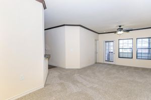 Spacious interior of a living area featuring light-colored walls, a ceiling fan, and large windows with horizontal blinds. The carpeted floor provides a cozy feel, and a door leads to an outdoor space. The area is well-lit and inviting, showcasing a modern design with a neutral color palette.