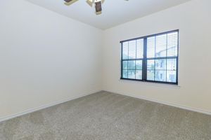 A vacant room with light beige walls and a ceiling fan. The floor is covered in gray carpet. There is a window with black blinds that provides a view of an outdoor area. The space is well-lit and appears clean, offering a simple and modern aesthetic.