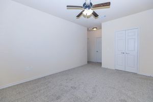 Empty room with light beige walls and carpet. A ceiling fan with wooden blades hangs from the ceiling. There's a closed door on the right and double doors leading to a closet. The room has no furniture and is well-lit from a ceiling light fixture.