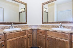 A spacious double sink bathroom with a brown wooden vanity, featuring two mirrors above the sinks. The countertops are made of a speckled granite material. The walls are a light, neutral color, and a door can be seen in the background. The overall design is modern and clean.