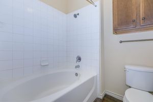 A clean and modern bathroom featuring a white tiled shower area, a built-in bathtub, and a toilet. Wood cabinets are visible above the tub, adding storage space. Soft lighting enhances the clean, spacious feel of the room.