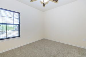 Empty room with beige walls and carpeted floor. A ceiling fan is mounted in the center, and a window with horizontal blinds allows natural light to enter. The room has no furniture and appears to be well-maintained. Light coming through the window adds to the spacious feel.