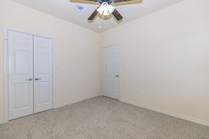 A small, empty room with beige walls and light gray carpet. It features a ceiling fan, two doors (one possibly a closet), and no furniture or decoration. The room has a simple and clean design, providing a blank space for potential use.