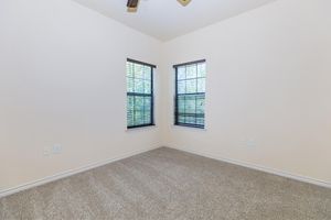 Empty room with light beige walls and carpet. Two windows on the left side feature dark wooden blinds, allowing natural light to filter in. The room has a ceiling fan and no furniture, creating a spacious and airy feel.