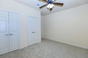 A light-colored room featuring beige walls and a carpeted floor. There are two white double doors on the left, and an additional door on the right. A ceiling fan with wooden blades is visible overhead, contributing to the room's spacious feel. The area is well-lit with no furniture present.
