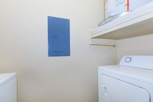 A laundry room featuring a stacked washer and dryer. The wall has a blue electrical panel and a shelf above the dryer, with pipes visible along the wall. The overall color scheme is light, creating a clean and functional space for laundry activities.