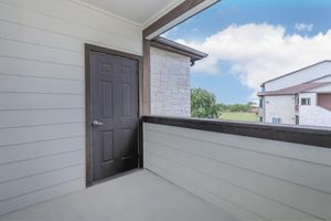 A view of a balcony featuring a dark door on the left side and a wooden railing. The walls are light-colored with a stone accent, and there’s a bright blue sky in the background. Other residential buildings can be seen in the distance. The scene is clean and spacious, ideal for outdoor relaxation.