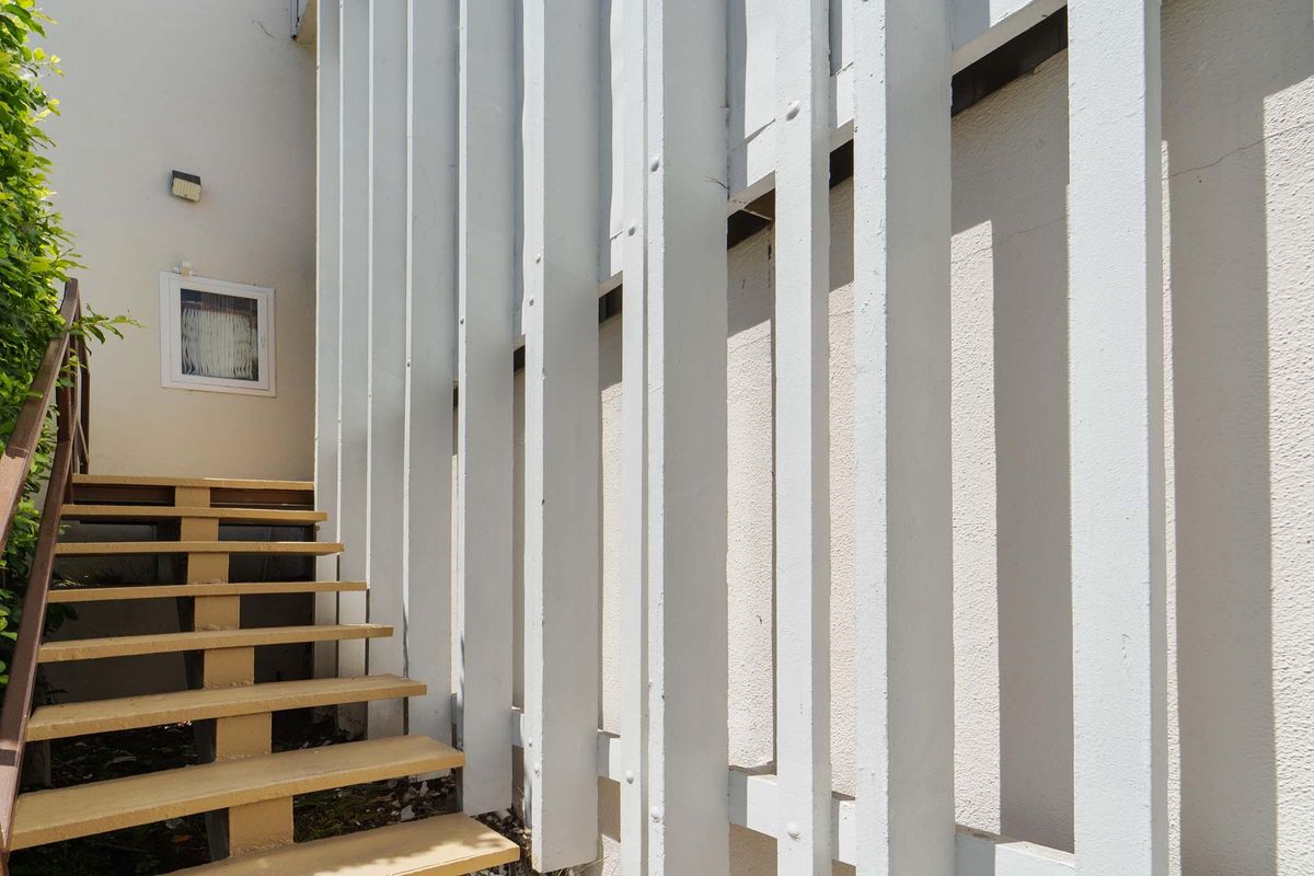 A staircase leading up alongside a wall with vertical white slats and a small window visible on the side. Natural light casts shadows on the stairs and wall.