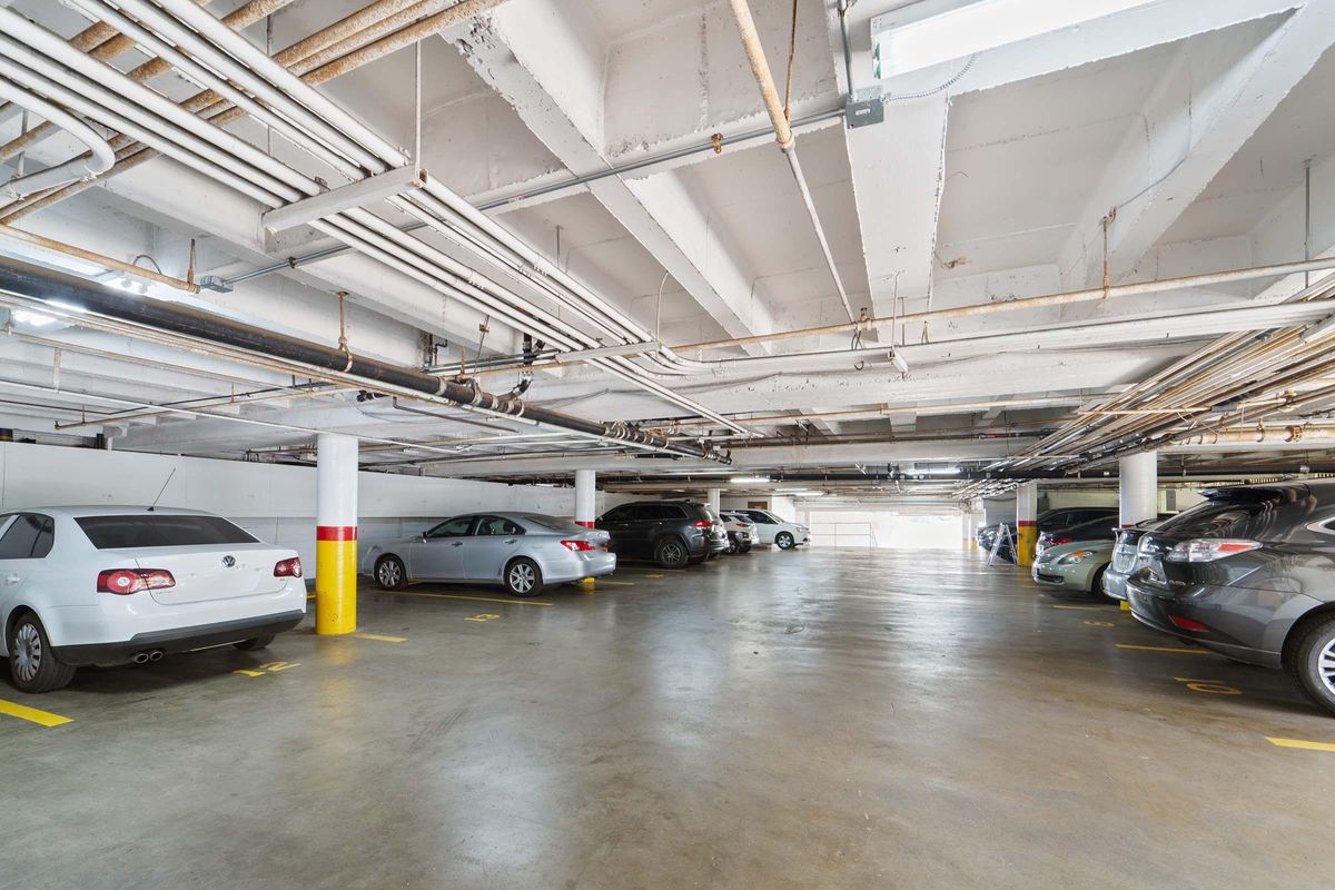 A spacious, well-lit underground parking garage with various parked cars and visible ceiling pipes. The concrete floor is marked with yellow parking lines, and there's a yellow-painted pillar in the foreground.