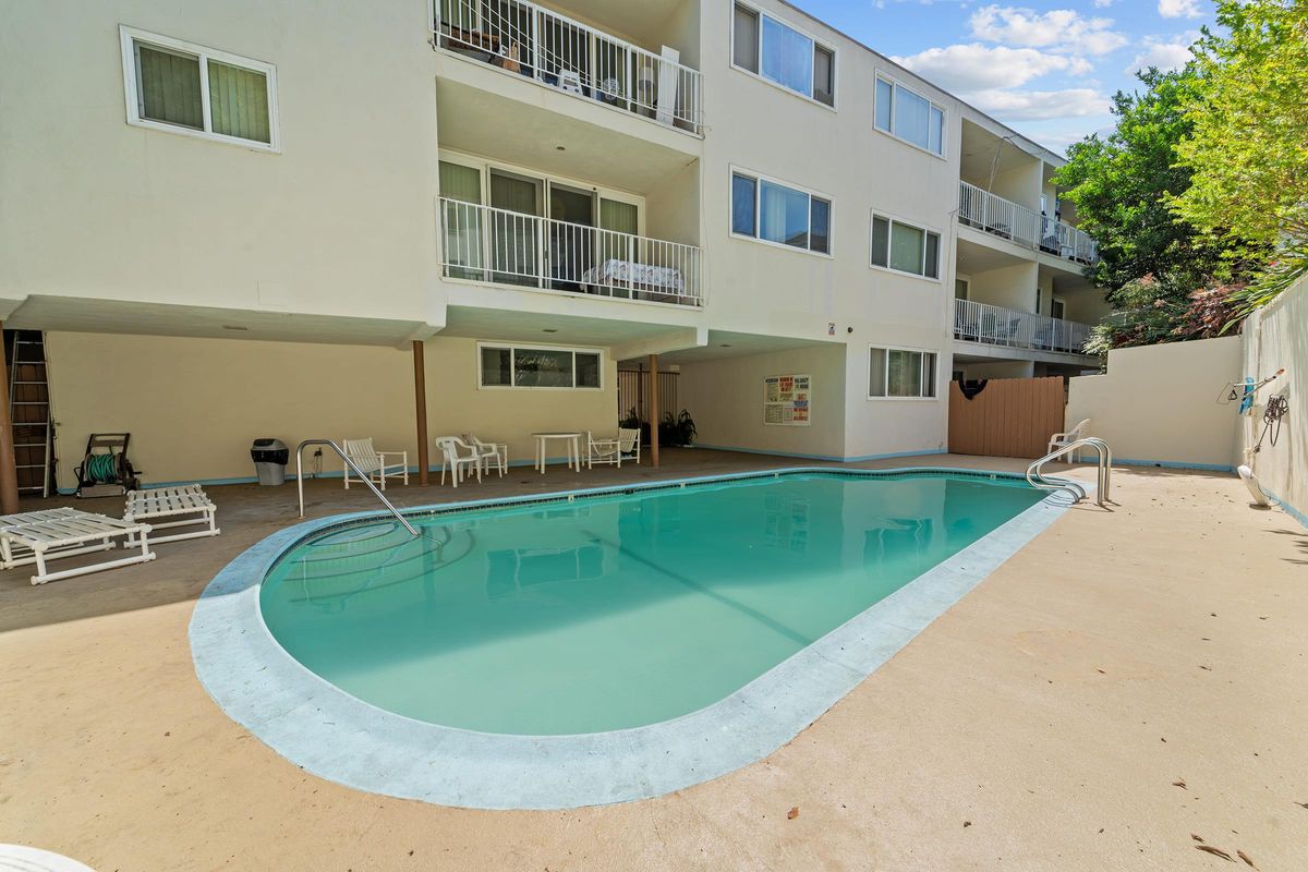 A clear swimming pool surrounded by a concrete deck, with lounge chairs positioned nearby. In the background, there is a multi-story building with balconies, and some greenery visible.