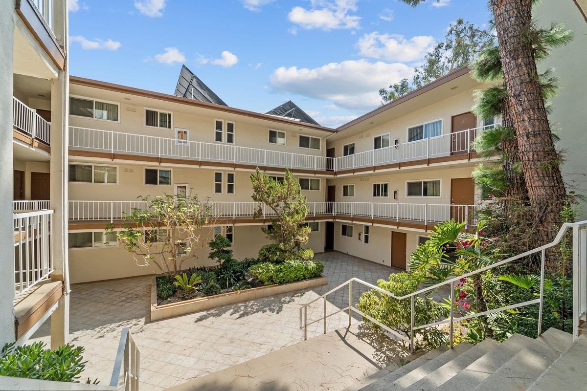 A view of an apartment courtyard featuring multiple levels of balconies, surrounded by greenery including trees and shrubs, under a partly cloudy sky.