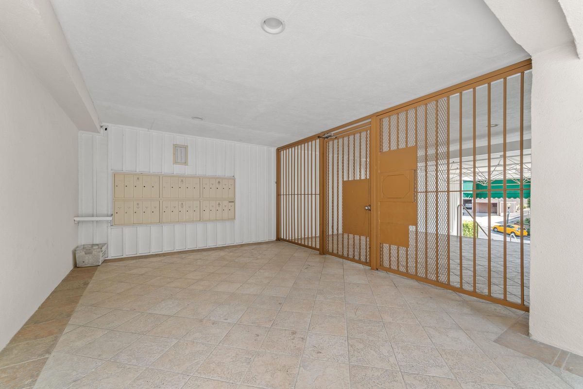 An interior entryway with a tiled floor and a gated entrance, featuring a row of mailbox slots on the left side against a white wall. The area is well-lit, with a ceiling light visible.