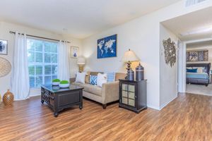 A cozy living room featuring a beige sofa adorned with decorative pillows, a coffee table, and a lamp. Large windows allow natural light to brighten the space. Wall art includes a world map, and a warm wooden floor enhances the inviting atmosphere. A doorway leads to another room in the background.