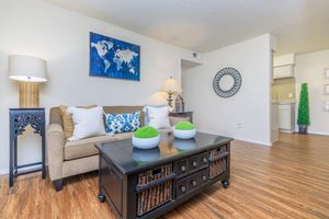 A cozy living room featuring a light brown sofa with decorative blue and white pillows, a dark coffee table with baskets, and a lamp on a decorative stand. The room includes a wall map art piece, a round mirror, and a glimpse of a kitchen area in the background. Green decorative plants add a touch of color.