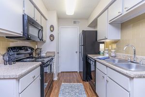 Modern kitchen with beige walls and light wooden flooring. Features include white cabinets, a black refrigerator, a black microwave, and an oven. There are stainless steel sinks, a decorative rug on the floor, and various kitchen utensils on the counter. A door leads to another room, adding accessibility.
